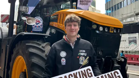 Jon Watt A head and torso shot of Jon Watt, standing in front of a large black tractor with an orange bonnet and orange-rimmed wheels. He has short mid-brown hair, curling on top, and is wearing a black zipped jacket with the NFU logo on it. He is holding a sign saying Back British Farming. 