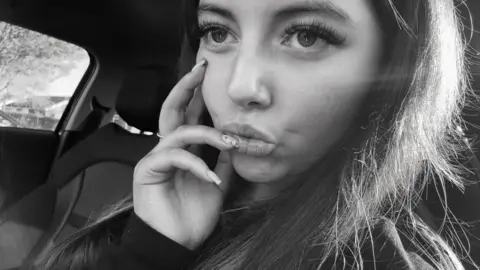 Black and white selfie of Sophie Claxton with long brown hair sitting in a car which her hand on her face and one nail resting on her lips. 