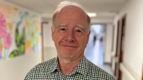 A man called George Oswald looks at the camera while standing in a hallway. He is wearing a green and black gingham shirt. A colourful mural is on the wall over his right shoulder.