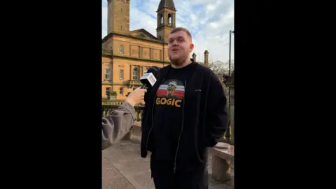 Man with short brown hair and graphic T-shirt and black jacket and jeans stands on the street and speaks into a mic.