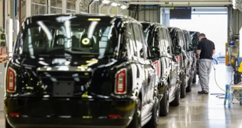 A row of black cabs lines up behind each other in a factory. There is a man in a black shirt and grey trousers holding a cable next to one of the cars.