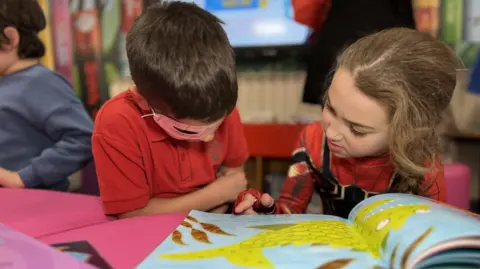 A young boy and girl are reading a book. The boy has dark hair and is wearing a Lone Ranger-style mask while the girl has light brown hair and has her finger paused over a word in the book.