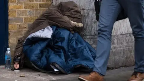 A homeless person sat on a street wearing a parka coat covering their face and wrapped in a sleeping bag with smartly dressed legs walking in front of them