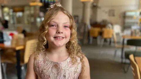 A little girl with blonde hair wearing a party dress smiles for the camera 