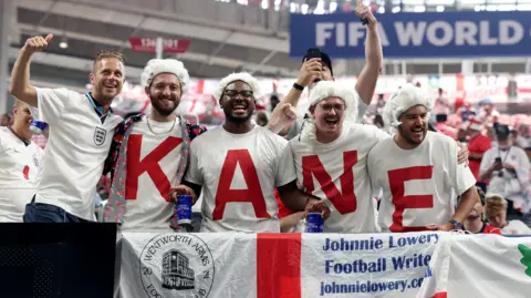 Reuters England fans display the name of Harry Kane on their shirts inside a stadium. They are all wearing white wigs.