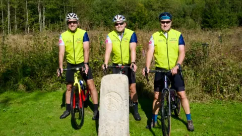 Three men with bicycles and wearing lime green gilets, beside a stone border post