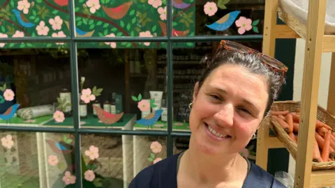 Ben Parker/BBC A woman wearing a blue top with sunglasses on her head, she is standing in front of a shop window with flower decals