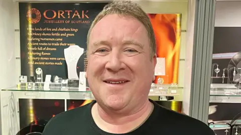 A man stands smiling in front of a display of Ortak jewellery, which includes broaches, necklaces, and earrings. He has blonde hair and is wearing a black top.