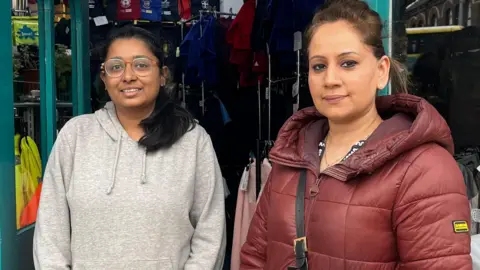Reena Fazil, who has brown hair, a red puffer coat and black handbag, smiles at the camera. On her right is her colleague, who has black hair, glasses and is wearing a grey hoody. 