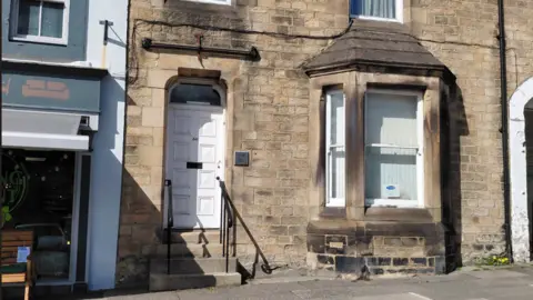 The register office in Barnard Castle is within a two-storey beige stone building with white windows. It has an arched doorway and a bay window.