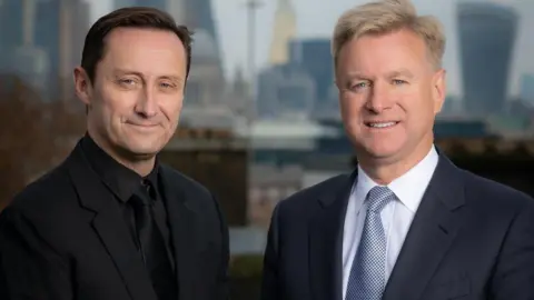 Involution of the Adam Beauty stands on the left. He has brown hair and wearing black coats and tie black. Tom is on the right and have a blonde hair with blue eyes. He wearing a dark armed armor in a white shirt with a blue-spotted tie. Both smiling into the camera.