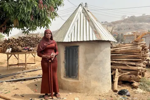 Adaobi Tricia Nwaubani Princess standing in front of her father’s traditional grave. She is wearing a long brown gown and standing next to a round concrete structure with a conical roof made from corrugate iron.