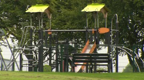 The play area in the park. There is a slide and climbing frames and a bench.