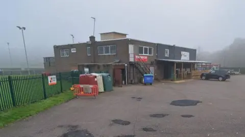 A two-storey brick sports clubhouse with flat roof, and a wooden awning on one side. Sport floodlights are visible on pitches beyond, with bins in the car park in front.