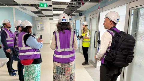 BBC/JULIA LEWIS A group of people in high visibility jackets stand in a hospital corridor