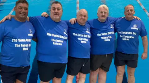 Five men pose arm in arm next to a swimming pool. They are wearing matching blue t-shirts with "The swim old gits" written on them and their names. All are looking at the camera.