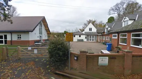 Google A primary school with a modern hall with white and red brick walls to the left, a yard in the middle and older brick buildings to the right. There is a white building with a triangular upper floor in the background, and a brick wall in the foreground.