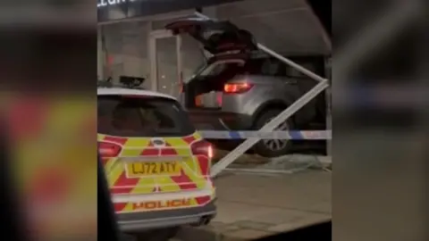 A grey car in the side of a building with the back of a police car in the corner of an image.