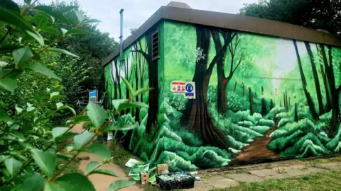 Anna Wheelhouse A green 3D forest with a trail leading through it, painted over a Cadent Gas building