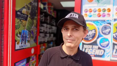 Vape manager Kamran Akhtar, wearing a black cap and black shirt, poses for a photo outside his vape shop