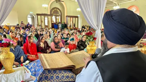 The picture is taken behind the priest. the book is in front of him and the crowd is in the background.