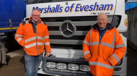 The two men are both wearing orange fluorescent jackets and trousers and are stood in front of a big lorry.