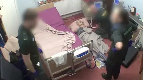 Kirstie Lumley A still from a CCTV camera shows a living room with a hospital bed in the middle of it. A 98-year old woman is lying on the floor and she is surrounded by paramedics.