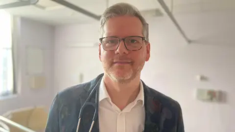 Simon Dedman/BBC A man wearing black framed glasses while looking at the camera. He is wearing a white buttoned shirt with the top button open. He is also wearing a black blazer and has a stethoscope around his neck 