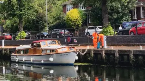 Owen Sennitt/BBC Anglian Water workers wearing orange high-vis protective clothing use a net to collect fish and put them in a blue bag, while stood along the bank of a river next to a motor cruiser