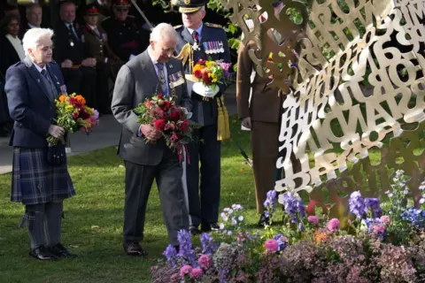 Getty Images King Charles III lays flowers at a brass memorial which is surrounded by a floral installation. He is flanked by two veterans who are carrying rainbow-coloured bouquets. The memorial is made of brass and resembles an unfolding letter made up of words given in evidence to a review of the treatment of LGBT+ Veterans. It is visible only in the top corner of the photograph.