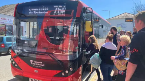 BBC A red 76A Transport for Cornwall bus is stationary on the side of the road and there is a line of passengers who are alighting the bus.