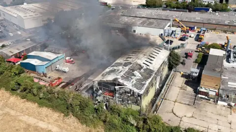 An aerial view of an industrial site. In the middle is a green metal building which is on fire. A large cloud of grey thick smoke is coming from the building. There are several cars around the site.