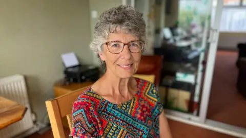 Woman with short grey curly hair and glasses and patterned blouse sat at wooden table in her living room