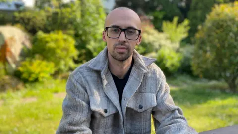 Myles is wearing a light grey jacket with a black t-shirt underneath. He is wearing black framed glasses and has a bald head with stubbly facial hair.