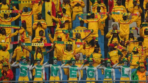 Seven football supporters stand and salute while spelling out the word Senegal with large letters painted on their bodies in green in front of numerous other Senegal fans who are predominantly wearing yellow and holding up Senegal scarves in the stand of a football stadium
