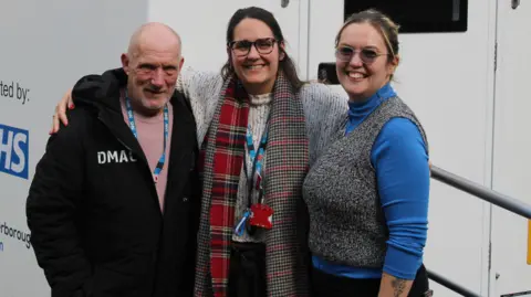 Light Project Peterborough Roy wearing a black coat standing next to Jess wearing a checked scarf and grey sweater and Heti wearing a blue jumper and a grey waistcoat, from the Boroughbury outreach team 