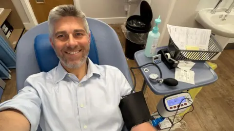 NHSBT Paul Bickley sits in a medical donation chair indoors with a blood pressure cuff and tubing attached to one arm, with blood donation equipment and a tray visible beside the chair.