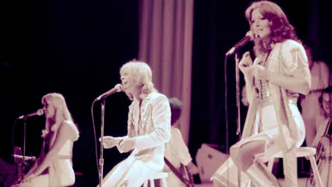 PA Media ABBA performing at the Royal Albert Hall in 1977. Three members of the band are sitting on stools in front of microphones singing.