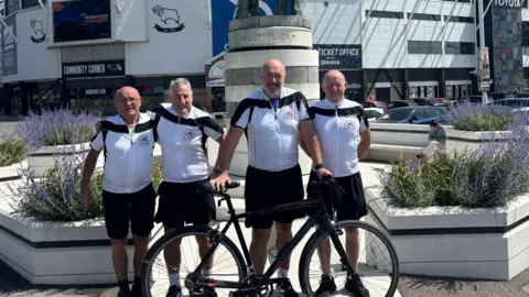 John Shiels. Cuatro hombres están de pie uno al lado del otro frente a la entrada de un club de fútbol. Uno de ellos sostiene una bicicleta negra. Todos visten camisetas blancas de ciclismo y pantalones cortos negros.