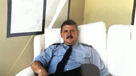 Avon Pensioners Jeff Lovell sitting on a white sofa in front of a beige wall. He is wearing a blue fire service shirt as part of his uniform, with black epaulettes on his shoulders. He looks to be about middle-age, has dark hair and a full moustache, and is looking at the camera.