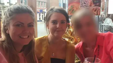 Facebook Three woman photographed in what appears to be a bar or a pub. The woman on the right is wearing a pink top and has had her face blurred. The two other women appear to be in their twenties, have dark hair and are smiling at the camera