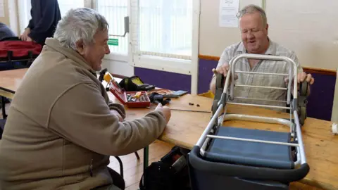 A man in a brown top with grey hair sits across a table from a man with a grey shirt. They are examining a white metal trolley with blue fabric.