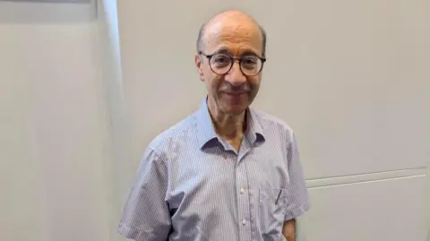 Paul Harris standing by a white wall, with short hair, glasses and wearing a stripped shirt.