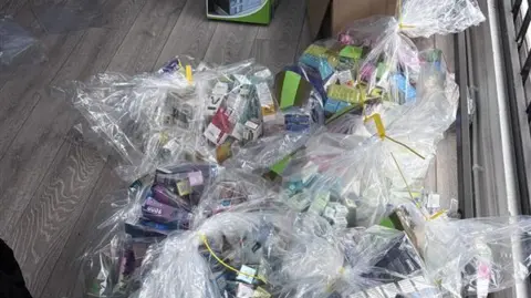 Clear plastic bags filled with colourful items, many in small cardboard boxes. They appear to be packets of vapes and cigarettes. They are coloured purple, green, blue and yellow. Yellow cable ties bind the plastic bags, which are sitting on a laminate wood floor.