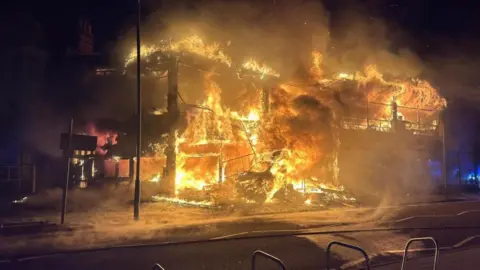 Essex County Fire and Rescue Service A building that is completely alight. Huge flames lick the frame of the building and there is a lot of smoke. It is night time.