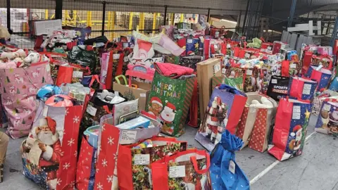 Charlotte Riley A large collection of colorful Christmas gift bags and boxes arranged on the floor in what appears to be a warehouse or distribution center. The bags feature festive designs such as Santa Claus, snowflakes, Christmas trees, and holiday greetings. Many of the bags are filled with toys, stuffed animals, and other gifts, and some have labels attached. 
