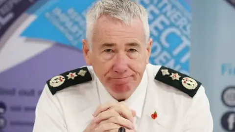 PA Media Jon Boutcher has grey hair and is leaning slightly forward in his PSNI shirt with his hands clasped in front of him