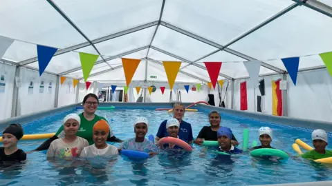 Children and adults stand in a large indoor swimming pool beneath a white tent canopy, decorated with colourful triangular bunting. Participants hold red, green, yellow and blue foam noodles and kickboards during a swimming session.