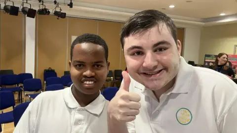 Abdul (left) a boy with dark hair, wearing a white t-shirt and Marc (right), a boy wearing a white t-shirt, who is holding his thumb up. You can see a yellow logo on Mark's shirt.