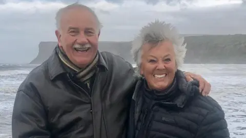 Trevor Sherman A smiling couple in their 70s stands in front of a seafront vista, with coastal and mountainous terrain in the distant background. The man is on the left, with grey hair and a moustache and dressed in a leather jacket, has arm around a woman with silver windswept hair and a puffer jacket on the right. 